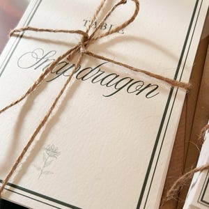 以下が含まれることがあります： 茶色の麻紐で結ばれたオフホワイトの長方形の紙カードの束。一番上のカードには、濃い緑色の二重線の枠、TABLEという文字、筆記体で書かれたSnapdragonという文字、そして下部に小さな花の線画が描かれています。