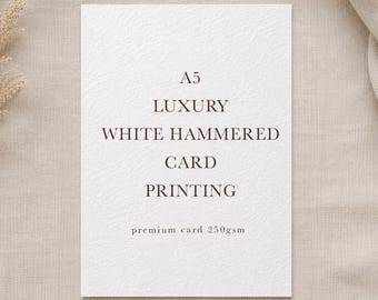 以下が含まれることがあります： ダークブラウンの文字で「A5 LUXURY WHITE HAMMERED CARD PRINTING premium card 250gsm」と書かれた、長方形のA5サイズの白いハンマートーン加工のカード。カードは質感のある表面を持ち、垂直方向に配置され、明確なタイポグラフィで表示されています。