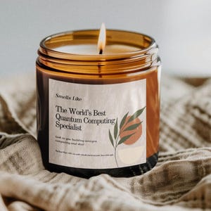 May include: A lit candle in a brown glass jar with a label that reads "The World's Best Quantum Computing Specialist." The label features a botanical design with a burnt orange and beige color scheme. The candle sits on a beige cloth.