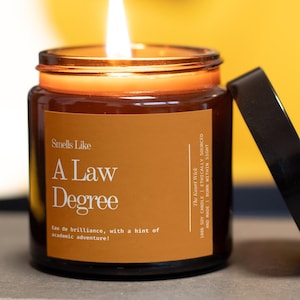 Puede incluir: Una vela encendida en un tarro de cristal marrón con tapa negra. La etiqueta dice "Smells Like A Law Degree". La vela está sobre un libro, con un fondo amarillo. La llama de la vela es brillante.