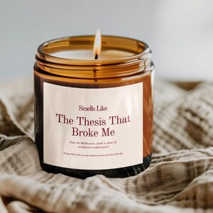Puede incluir: Una vela encendida en un tarro de cristal marr&oacute;n. La etiqueta dice "Smells Like The Thesis That Broke Me". La vela est&aacute; sobre una tela beige. La llama de la vela est&aacute; encendida y el tarro tiene un borde dorado.