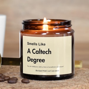 May include: A lit candle in an amber glass jar with a cream label reading "Smells Like A Caltech Degree". Next to the candle is a gold-toned metal measuring scoop containing dark roasted coffee beans, with a white mug in the background.