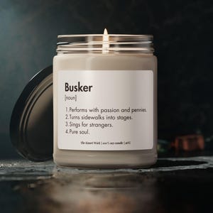 Può includere: Una candela accesa color crema in un barattolo di vetro con coperchio argentato. L'etichetta recita "Busker" con una definizione. La candela è su una superficie scura, con il coperchio leggermente socchiuso. Lo sfondo è blu scuro.