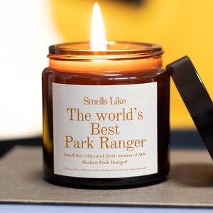 May include: A lit, brown glass candle jar with a black lid. The label reads "Smells Like The world's Best Park Ranger." The candle is on a grey surface with a yellow background. The candle's flame is bright.