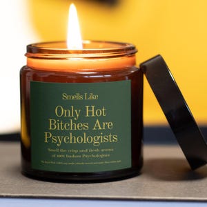 May include: A lit, brown glass jar candle with a black lid. The candle has a green label with the text "Smells Like Only Hot Bitches Are Psychologists". The candle is on a neutral surface with a yellow background.
