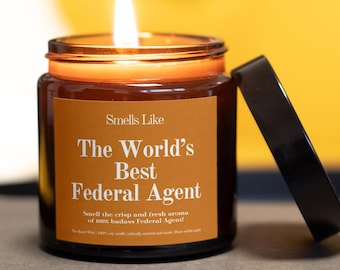 May include: A lit, amber-colored glass candle jar with a black lid. The label reads "Smells Like The World's Best Federal Agent." The candle is on a neutral surface with a yellow background.