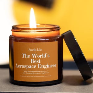 May include: A lit, brown glass jar candle with a black lid. The candle has a label that reads "Smells Like The World's Best Aerospace Engineer." The candle is on a neutral surface with a yellow background.
