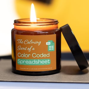 May include: A lit candle in a brown glass jar with a black lid. The label reads "The Calming Scent of a Color Coded Spreadsheet" with an Excel icon. The candle is on a neutral surface with a yellow background.
