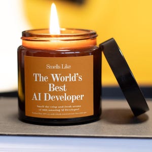 Puede incluir: Una vela encendida en un tarro de cristal ámbar con tapa negra. La etiqueta de la vela dice "Smells Like The World's Best AI Developer". La vela está sobre una superficie neutra con un fondo amarillo.