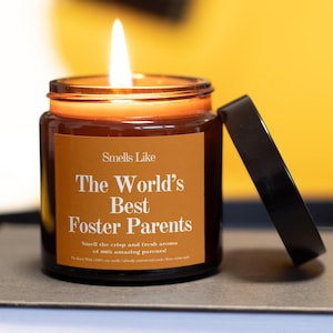 Peut inclure: Une bougie en pot de verre brun allumée avec un couvercle noir. La bougie porte une étiquette indiquant "Smells Like The World's Best Foster Parents". La bougie est sur une surface grise avec un fond jaune.