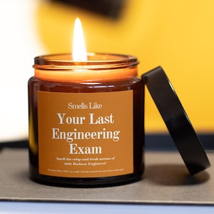 May include: A lit candle in a glass jar with a brown label that reads "Smells Like Your Last Engineering Exam". The black lid is resting next to the jar. The candle is on a stack of books.