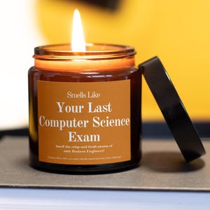 Puede incluir: Una vela de vidrio marrón encendida con una tapa negra. La vela tiene una etiqueta que dice "Smells Like Your Last Computer Science Exam". El texto debajo dice "Smell the crisp and fresh aroma of 100% Badass Engineer!"