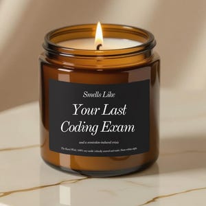 Może przedstawiać: Zapalone brązowe szklane naczynie na świece z czarną etykietą. Na etykiecie widnieje napis "Smells Like Your Last Coding Exam" białym tekstem. Świeca stoi na białej i złotej powierzchni.