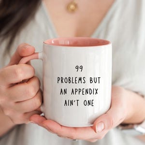 Puede incluir: Taza de cerámica blanca con interior y asa rosas. La taza presenta el texto negro "99 PROBLEMS BUT AN APPENDIX AIN'T ONE" centrado en la parte delantera. La taza es sostenida por una persona.