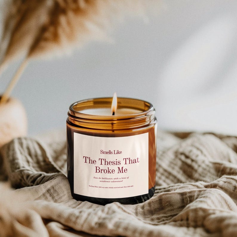 Puede incluir: Una vela encendida en un tarro de cristal marr&oacute;n. La etiqueta dice "Smells Like The Thesis That Broke Me". La vela est&aacute; sobre una tela beige. La llama de la vela est&aacute; encendida y el tarro tiene un borde dorado.