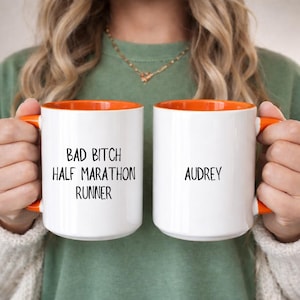 May include: Two white ceramic mugs with orange interiors and handles. One mug reads "BAD BITCH HALF MARATHON RUNNER" in black text. The other mug has the name "Audrey" in black text. The mugs are held by a person.