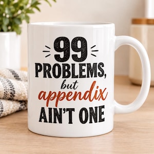 Op de afbeelding: Een witte keramische mok met zwarte en oranje tekst die 99 PROBLEMS, but appendix AIN'T ONE leest. De mok heeft een standaard handvat en een gladde, glanzende afwerking, waarbij de donkere letters duidelijk op het witte oppervlak staan.