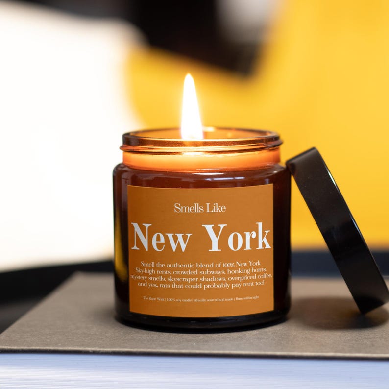 May include: A lit candle in a glass jar with a brown label that reads "Smells Like New York." The text below describes the scent. The lid is off to the side. The candle sits on a book with a yellow background.