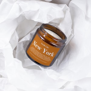 May include: A brown glass jar candle with a white wax interior, labeled "New York" in white text. The candle sits on a bed of white tissue paper. The label also includes the text "Smells Like".