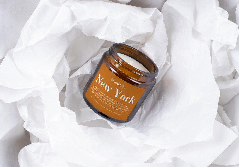 May include: A brown glass jar candle with a white wax interior, labeled "New York" in white text. The candle sits on a bed of white tissue paper. The label also includes the text "Smells Like".