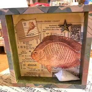 May include: A vintage wooden shadow box with a collage of paper ephemera, including a fish, seashells, and a star. The box has a green and brown camouflage pattern on the outside. The text on the paper ephemera includes "Islands-IVORY COAST", "Garibaldi I", "Types of Italian Stamps", "Overprinted in Red or Blue", "10c brown", "20c red brown (BI)", "Ferrucci I", "Types of Italian Stamps", "Overprinted in Red or Blue", "shine", "15°", "L.H.", and "POSTES AFR FRANCAN".
