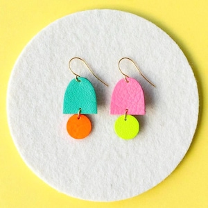 以下が含まれることがあります： ターコイズ、ピンク、オレンジ、イエローのカラーリングのカラフルな幾何学模様のイヤリングのペア。イヤリングは革製で、ゴールドトーンの仕上げです。