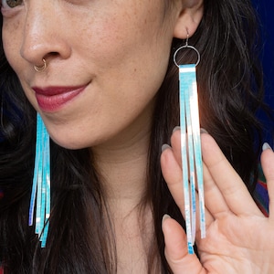 May include: A pair of long, dangling earrings featuring a circular silver-toned metal hoop at the top. Attached to each hoop are multiple thin, vertical strips of iridescent, light-reflecting material that shimmer with shades of blue and white.