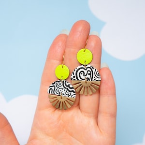 May include: A pair of dangle earrings with a black and white swirl pattern on a white background. The earrings have a gold-colored sunburst design and a bright yellow circle at the top.