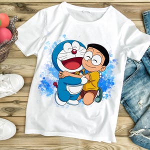 May include: White t-shirt featuring a cartoon illustration of Doraemon and Nobita hugging against a blue watercolor splash background. The characters are in blue, yellow, and brown. The shirt is on a wooden surface.