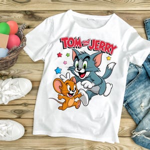May include: White t-shirt featuring the cartoon characters Tom and Jerry. The graphic shows Tom the cat in blue and Jerry the mouse in orange, with the words "Tom and Jerry" in red. Colorful stars surround the characters.