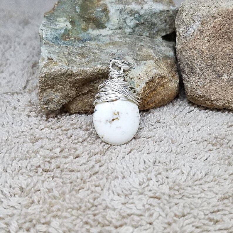 Puede incluir: Un colgante de piedra ovalada blanca envuelto en alambre plateado. La piedra tiene un patr&oacute;n moteado de peque&ntilde;as motas doradas.