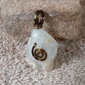 Puede incluir: Un colgante de piedra blanca opaca de forma irregular con un diseño en espiral de alambre de latón. El colgante está unido a un bucle de alambre de latón.