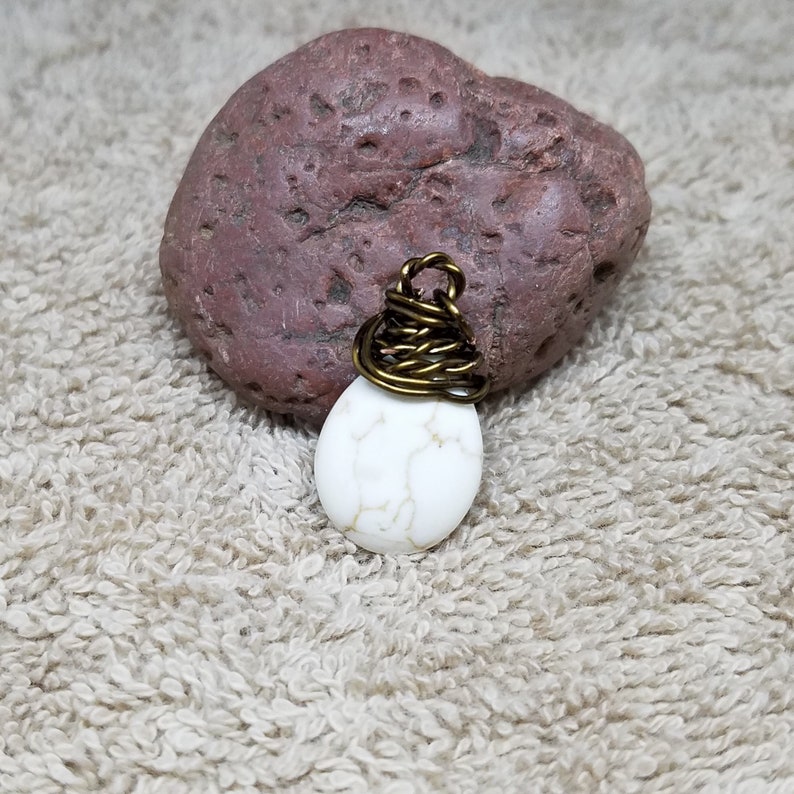 Puede incluir: Un colgante de piedra howlite blanca con un envoltorio de alambre de lat&oacute;n. El colgante est&aacute; sobre una superficie marr&oacute;n texturizada.
