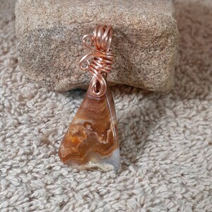 Puede incluir: Un colgante artesanal con una piedra triangular pulida con patrones marrones y blancos. La piedra está envuelta en alambre de cobre, formando una fianza decorativa. El colgante se muestra sobre una superficie de color neutro.