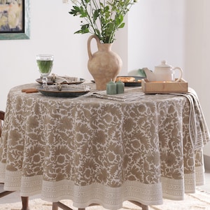 Op de afbeelding: Een ronde tafel is bedekt met een tafelkleed met bloemenpatroon in beige en wit. Een vaas met witte, roze en groene bloemen staat in het midden. De tafel is gedekt met borden, servetten en een theepot op een houten dienblad.