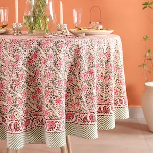 May include: A round tablecloth with a floral pattern in pink and green, set on a wooden table. The table is set with candles, glassware, and a vase of flowers. The tablecloth has a striped border.
