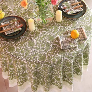 May include: A round tablecloth with a green and white floral pattern covers a table set for a meal. Green plates, napkins, and cutlery are arranged on the table. Candles and a vase of flowers add to the decor. An orange sits on a napkin.