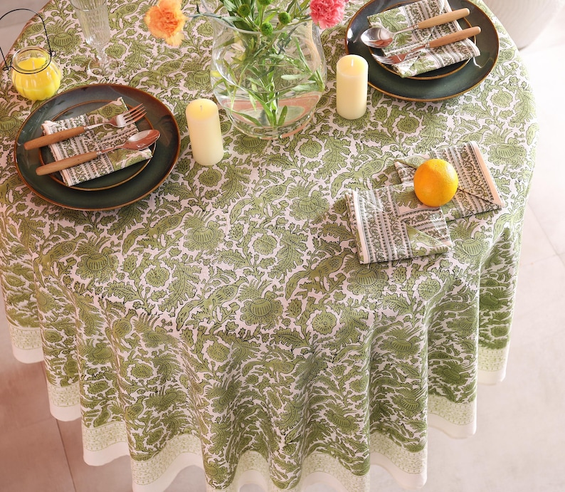 May include: A round tablecloth with a green and white floral pattern covers a table set for a meal. Green plates, napkins, and cutlery are arranged on the table. Candles and a vase of flowers add to the decor. An orange sits on a napkin.