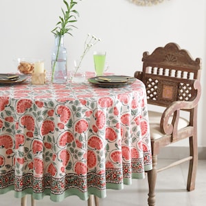 May include: A round tablecloth with a floral pattern in red and green on a cream background. The tablecloth is set on a wooden table with a wooden chair. A vase with flowers and a glass of green liquid are on the table.