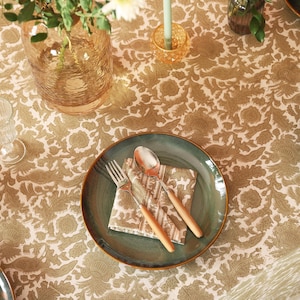 Puede incluir: Un plato verde azulado con borde dorado contiene una servilleta doblada, un tenedor y una cuchara con mangos de madera. La mesa está cubierta con un mantel con estampado floral en tonos beige y verde oliva. Un jarrón y un candelabro también están sobre la mesa.