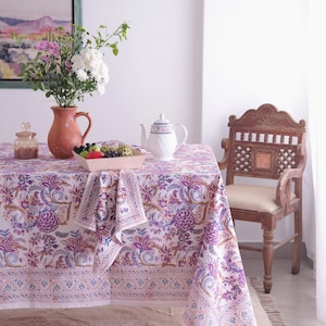 May include: A dining table set with a floral patterned tablecloth in shades of purple, white, and brown. A matching napkin is on the table. A teapot, vase of flowers, and a wooden bowl of fruit are on the table.