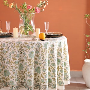 May include: A round tablecloth with a floral pattern in shades of green and blue, set for a meal. The table is adorned with a clear vase of flowers, candles, plates, and glasses. An orange sits on a folded napkin.
