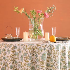 Puede incluir: Una mesa puesta para una comida, con un mantel blanco con un estampado floral verde. La mesa est&aacute; decorada con platos, velas, vasos y un jarr&oacute;n con flores. Una naranja est&aacute; colocada sobre la mesa. El fondo es de color naranja.