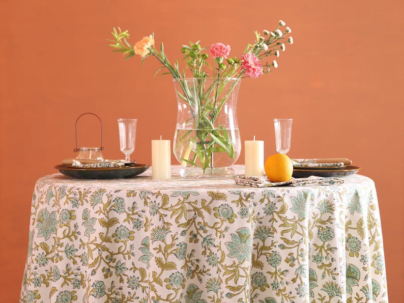 Puede incluir: Una mesa puesta para una comida, con un mantel blanco con un estampado floral verde. La mesa est&aacute; decorada con platos, velas, vasos y un jarr&oacute;n con flores. Una naranja est&aacute; colocada sobre la mesa. El fondo es de color naranja.