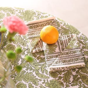 May include: A still life composition showing a folded napkin with a white and green pattern, an orange, and a matching tablecloth. Pink flowers and green buds are in the foreground. The tablecloth and napkins have a detailed floral design.
