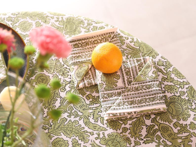 May include: A still life composition showing a folded napkin with a white and green pattern, an orange, and a matching tablecloth. Pink flowers and green buds are in the foreground. The tablecloth and napkins have a detailed floral design.