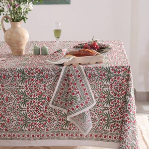 Op de afbeelding: Een eettafel gedekt met een bloemmotief tafelkleed en bijpassende servetten. Het witte tafelkleed heeft een patroon van rode bloemen en groene bladeren. De tafel is gedekt met glazen, kaarsen en een houten dienblad met brood.