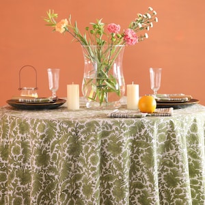 May include: A round table set for a meal, covered with a green and white floral tablecloth. The table is set with plates, napkins, candles, and a vase of flowers. An orange sits on the table.