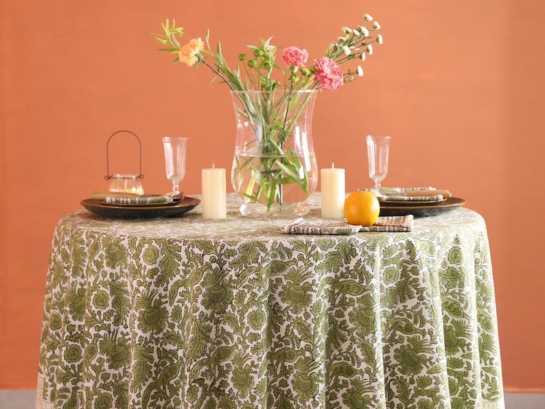 May include: A round table set for a meal, covered with a green and white floral tablecloth. The table is set with plates, napkins, candles, and a vase of flowers. An orange sits on the table.