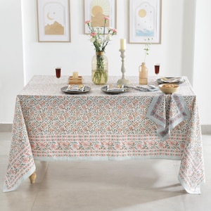 Op de afbeelding: Een witte tafelkleed met een roze en groen bloemenpatroon. De tafel is gedekt met borden, servetten en een kom met eten. Decoratieve items zijn een vaas met bloemen, kaarsen en ingelijste kunstwerken.